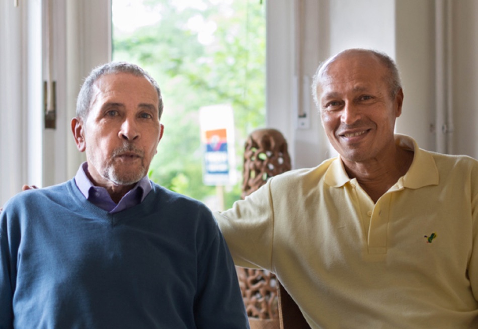 Two older men sitting side by side by a window, one in a blue sweater and one in a yellow polo shirt
